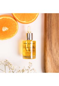 Żółty serum w szklanej butelce z zakraplaczem i srebrną nakrętką, otoczone połówkami pomarańczy, białymi kwiatami i drewnianą deską.