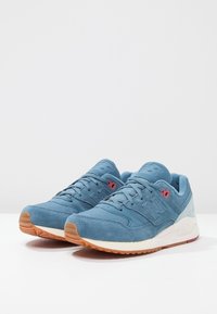 Paire de baskets New Balance en daim bleu avec semelles en gomme et intermédiaires crème sur une surface blanche contre un fond uni.