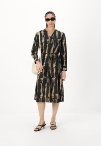 Juodas ir auksinis raštuotas suknelė su V formos iškirpte, ilgomis rankovėmis ir pririšta juosta. Pora su juodais sandalais su dirželiais ir šviesia rankine.