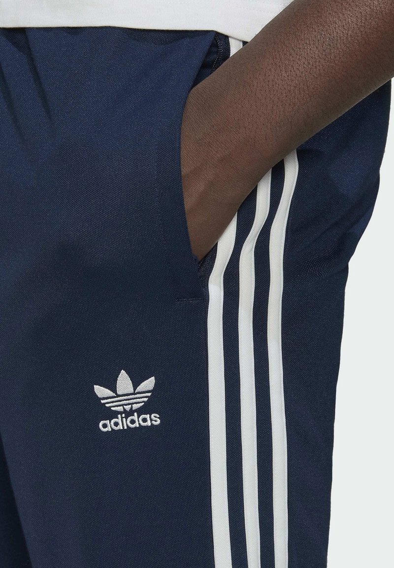 Pantalones deportivos azul marino con tres franjas blancas a lo largo del costado y un logotipo de Adidas bordado en blanco en el muslo izquierdo.