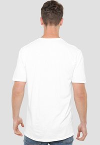 A person is seen from behind, wearing a plain white t-shirt and blue jeans, against a neutral gray background.