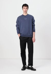 Felpa in cotone blu navy con polsini e orlo a costine, abbinata a pantaloni neri e scarpe nere, su uno sfondo semplice. Design e vestibilità semplice.