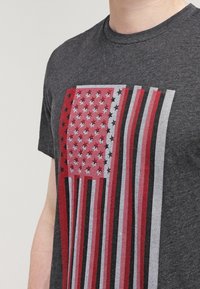 Camiseta de color gris oscuro con un gráfico en rojo, blanco y azul que se asemeja a la bandera americana, con detalles de rayas y un patrón de estrellas.