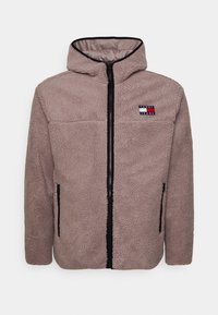 Chaqueta de forro polar marrón claro con cremallera y capucha. Presenta ribetes negros y un pequeño logotipo en el lado izquierdo del pecho. Tiene dos bolsillos laterales.
