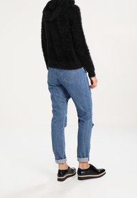 Schwarzer, flauschiger Kapuzenpullover, hellblaue Jeans mit umgeschlagenen Säumen und schwarze Schnürschuhe mit dicker Sohle, die ein lässiges Outfit von hinten zeigen.