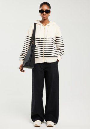 Žena koja nosi crne sunčane naočale, kremasti i crni prugasti duks na patentni zatvarač, široke crne hlače, bijele tenisice i nosi crnu torbu za nošenje.