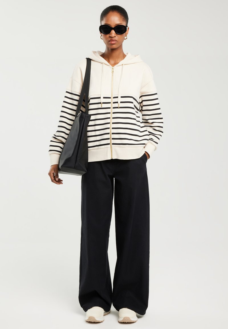 Kvinde iført sorte solbriller, creme- og sortstribet hættetrøje med lynlås, vide sorte bukser, hvide sneakers og bærer en sort taske.