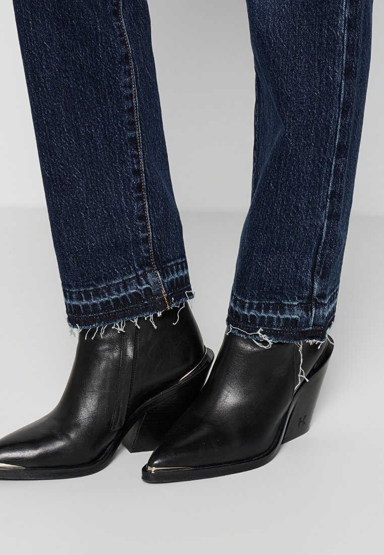 Bottines noires à talons avec des bouts pointus, associées à un jean en denim bleu foncé avec des ourlets effilochés et des coutures contrastées.