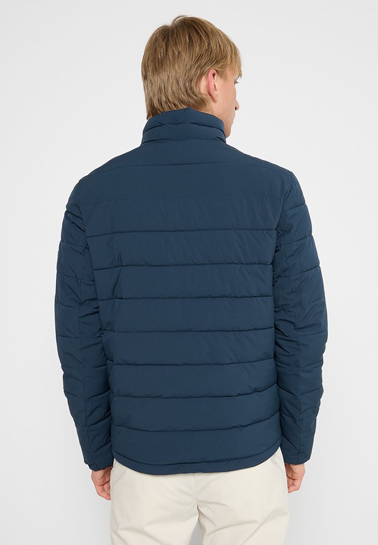 Man die met zijn rug naar de camera staat, gekleed in een marineblauwe gewatteerde jas en lichtbeige broek, tegen een plain witte achtergrond.