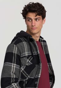Kurtka w kratę w kolorach czarnym, szarym i białym, z kapturem. Posiada zapinane na guziki kieszenie oraz bordową koszulę pod spodem. Teksturowana tkanina.