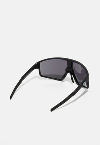 Czarne okulary przeciwsłoneczne w stylu owiewającym, z ciemnymi soczewkami i teksturowanymi zausznikami. Zawierają logo po boku. Lekki i sportowy styl.