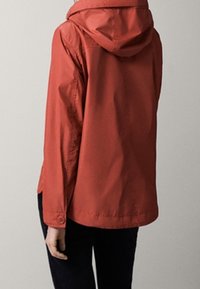 Person som bär en rostfärgad huvtröja med långa ärmar och knäppta manschetter, sedd bakifrån mot en enfärgad bakgrund.