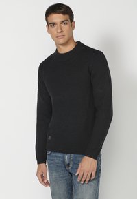 Dunkelblauer Strickpullover mit einem strukturierten Muster, Rundhalsausschnitt und gerippten Bündchen. Getragen über hellblauen Jeans, die ein kleines Markenlabel aufweisen.
