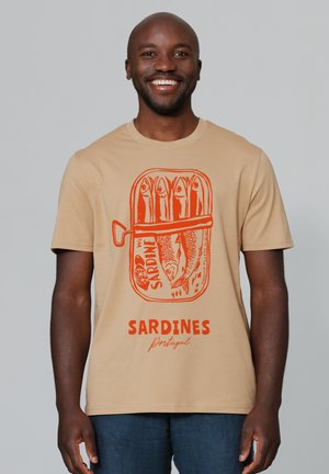 T-shirt beige avec un graphique de boîte de sardines orange, comprenant des illustrations de poissons et le texte "SARDINES Portugal" bien en vue.