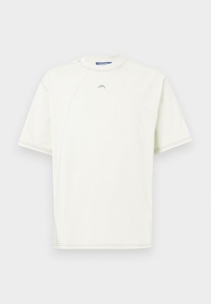 Vaalean kerma sydänhihainen T-paita, joka on valmistettu puuvillasta. Tässä paidassa on raglan-hihat, pyöreä kaula-aukko ja minimaalista ompelu yksityiskohtia. Pienen logo edessä.