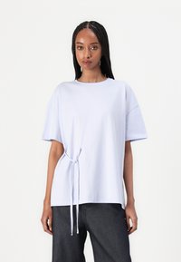 Lys lilla t-skjorte med rund hals, korte ermer og sidesløyfe. Stoffet ser glatt ut, løs passform med et enkelt design.