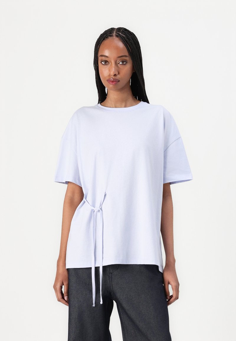 Lys lilla t-skjorte med rund hals, korte ermer og sidesløyfe. Stoffet ser glatt ut, løs passform med et enkelt design.