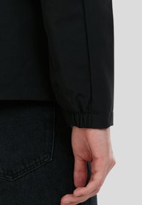 Sort, let jACKET med elastiske manchetter, glat tekstur, og et n�ærbillede af en hånd der hviler mod en mørk denim baggrund.