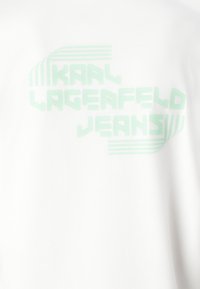 Valkoinen T-paita, jossa on mintunvihreä teksti "KAAL LAGERFELD JEANS" geometrisella fontilla, jossa on vaakasuoria viivoja ja sileä pinta.