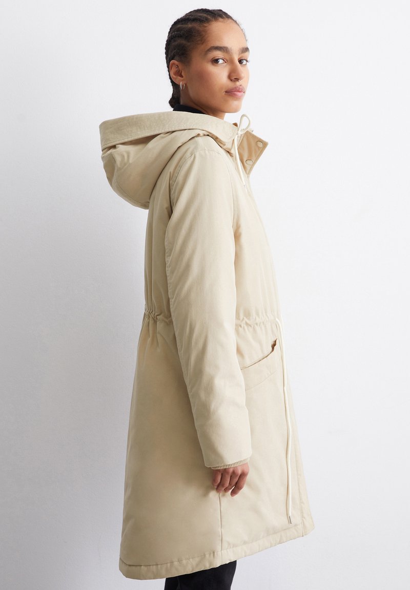 Beige gepolsterter Parka mit Kapuze, Kordelzug in der Taille, seitlichen Taschen und gerippten Bündchen. Glatte Textur, knielanges Design.