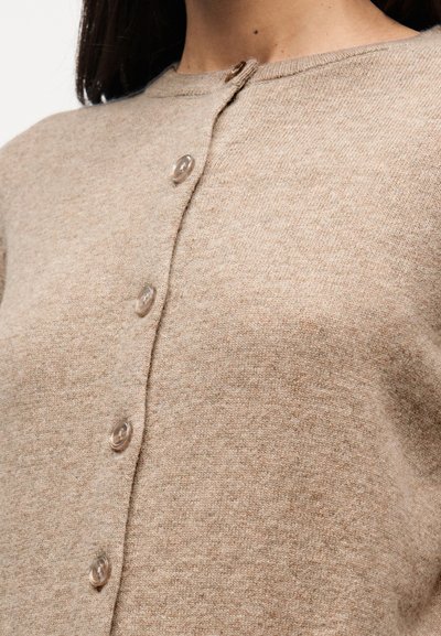 Μπεζ ζακέτα με στρογγυλή λαιμόκοψη, υφασμάτινη υφή και διαφανή κουμπιά μπροστά. Κοντινή λήψη που αναδεικνύει το υλικό και το σχέδιο.