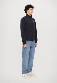Chaqueta de navy con cremallera y cuello alto, con puños y dobladillo elásticos. Combinada con jeans de un azul claro y zapatillas blancas.
