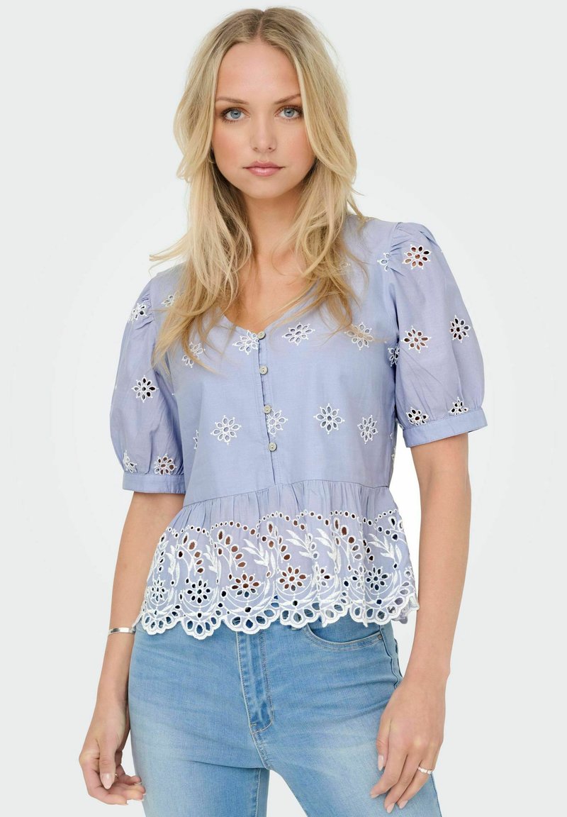 Blouse bleu clair avec des manches bouffantes et un devant à boutons, ornée de broderies florales et de dentelle le long de l'ourlet. Tissu en coton texturé.