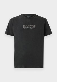 Camiseta negra de manga corta con el texto "GUESS Desde 1981 Diseño Auténtico" y elementos decorativos impresos en el pecho.