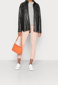 Persona que lleva una chaqueta de cuero negra, una camisa blanca y negra a rayas, pantalones rosa claro, zapatos blancos, y sostiene un pequeño bolso acolchado naranja.