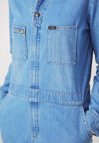 Combinaison en denim bleu clair avec poches sur la poitrine, une poche zippée à gauche et un bouton avec le logo "Lee".
