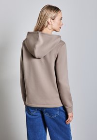 Felpa con cappuccio marrone dal texture morbido, caratterizzata da una tasca a canguro, polsini e orlo rigati, abbinata a jeans di denim blu.