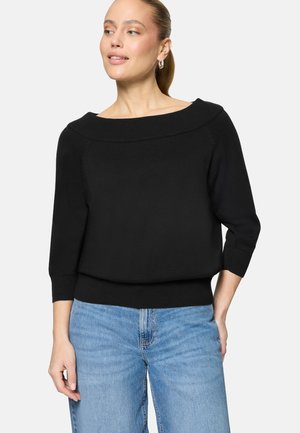 Femme à la peau claire et aux cheveux attachés porte un pull noir à col bateau et un jean bleu clair, regardant légèrement sur le côté, souriant doucement.