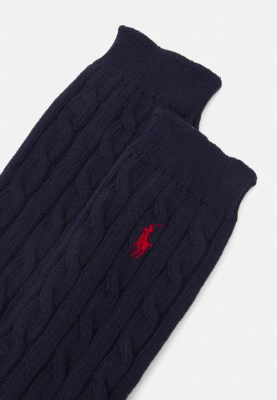 Jūras zilas adītas rokas sildītājas ar kabelīšu rakstu, kurām ir sarkans izšūts logo. Mīksta tekstūra ar ribotiem malām.