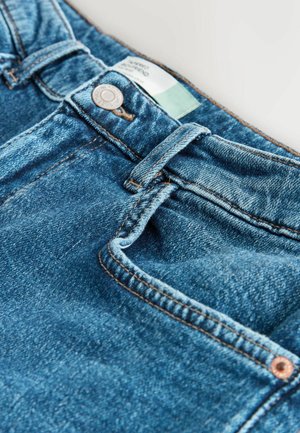Gros plan sur un jean en denim bleu montrant le bouton, les passants de ceinture, la poche avant, les coutures et une partie de l'étiquette de la marque à l'intérieur de la ceinture.