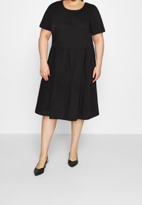 Schwarzes Kurzarmkleid mit Rundhalsausschnitt, gestuftem Design und lockerer Passform. Der Stoff wirkt glatt und leicht. Trägt dazu spitze schwarze Absätze.