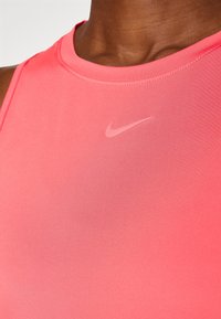 Rů�žový sportovní tílko s kulatým výstřihem a tenkými ramínky; má jemné logo Nike v světlejší barvě. Hladká textura a přiléhavý střih.
