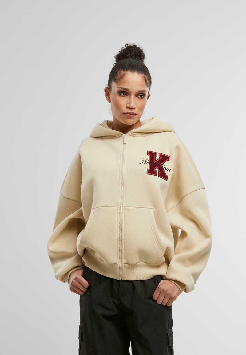 Bež zip-hoodie s velikim crvenim izvezenim slovom "K" na lijevoj strani prsa, oversize kroj, bočni džepovi, izrađen od mekanog, teksturiranog materijala.