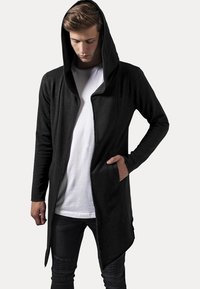 Sudadera negra larga con cremallera frontal, capucha grande y bolsillos laterales; fabricada con una tela suave de textura lisa.