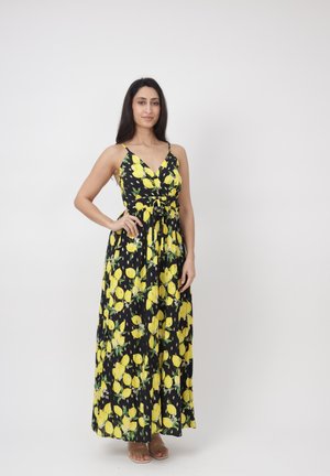 Kobieta stojąca z ręką na biodrze, ubrana w czarną bezrękawową sukienkę maxi w żółte cytrynowe wzory oraz beżowe sandały na białym tle.