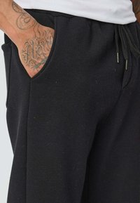 Un pantalon de survêtement noir avec un cordon de serrage à la taille, une texture de tissu douce, des poches latérales et une coupe ajustée, avec un tatouage visible sur la main.