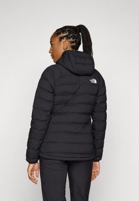 Svart pufferjacka med en figurnära design, huva och kviltad textur. Har "The North Face"-logotypen på övre delen av ryggen.