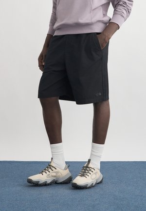 Person, die schwarze knielange Shorts, weiße Socken, beige-graue Sneakers und einen hellvioletten Pullover trägt und auf einem blauen Teppich steht.