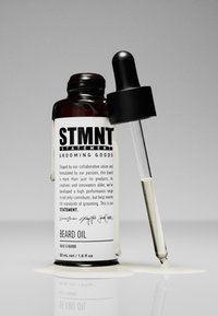 Brun glasflaska med skäggolja, med en svart droppare och en vit etikett med produktinformation. En oljespill under flaskan.