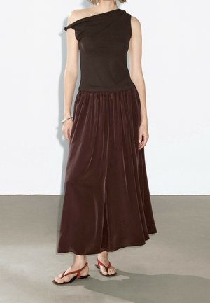 Femme portant un top asymétrique marron foncé à une épaule et une longue jupe fluide assortie avec des sandales rouges, debout contre un mur blanc.