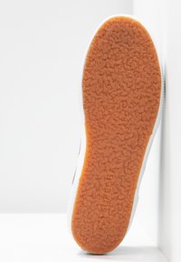 Semelle texturée marron d'une basket blanche, inclinée contre un mur d'angle blanc et une surface de sol blanche.