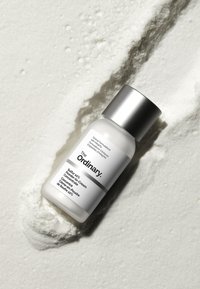 Witte, cilindrische fles met een zilveren dop. Het etiket heeft vet, zwart tekst. Omringd door een gestructureerd, poederachtig wit materiaal.