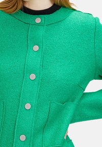Grön texturerad kofta med rund halsringning, silverfärgade tryckknappar och framfickor. Tyget har ett ludet utseende.