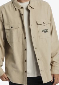 Beige flannel shirt with two front pockets, button closure, and embroidered logo. Made of textured fabric with a relaxed fit and brown buttons.