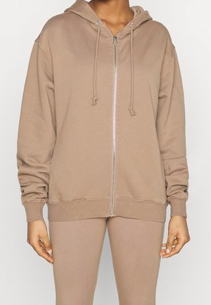 Personne portant un sweat à capuche beige zippé avec des cordons et des leggings beige assortis, debout devant un fond uni.
