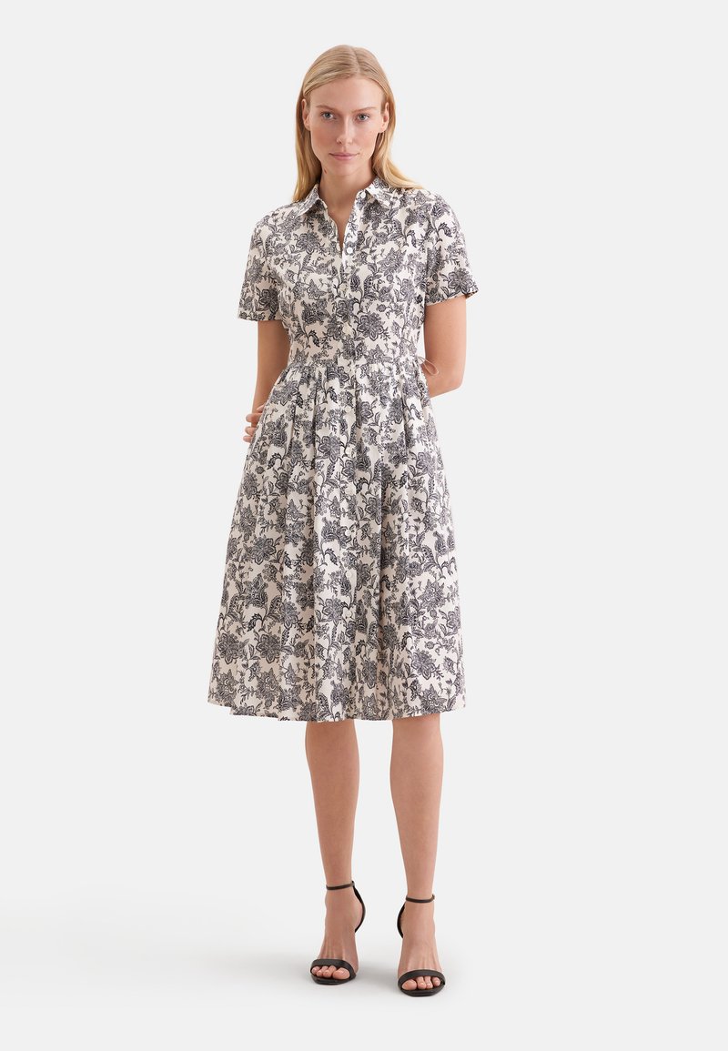 Femme portant une robe blanche longueur genou avec des motifs floraux noirs, manches courtes, col et talons noirs à bride à la cheville, debout les mains dans le dos.